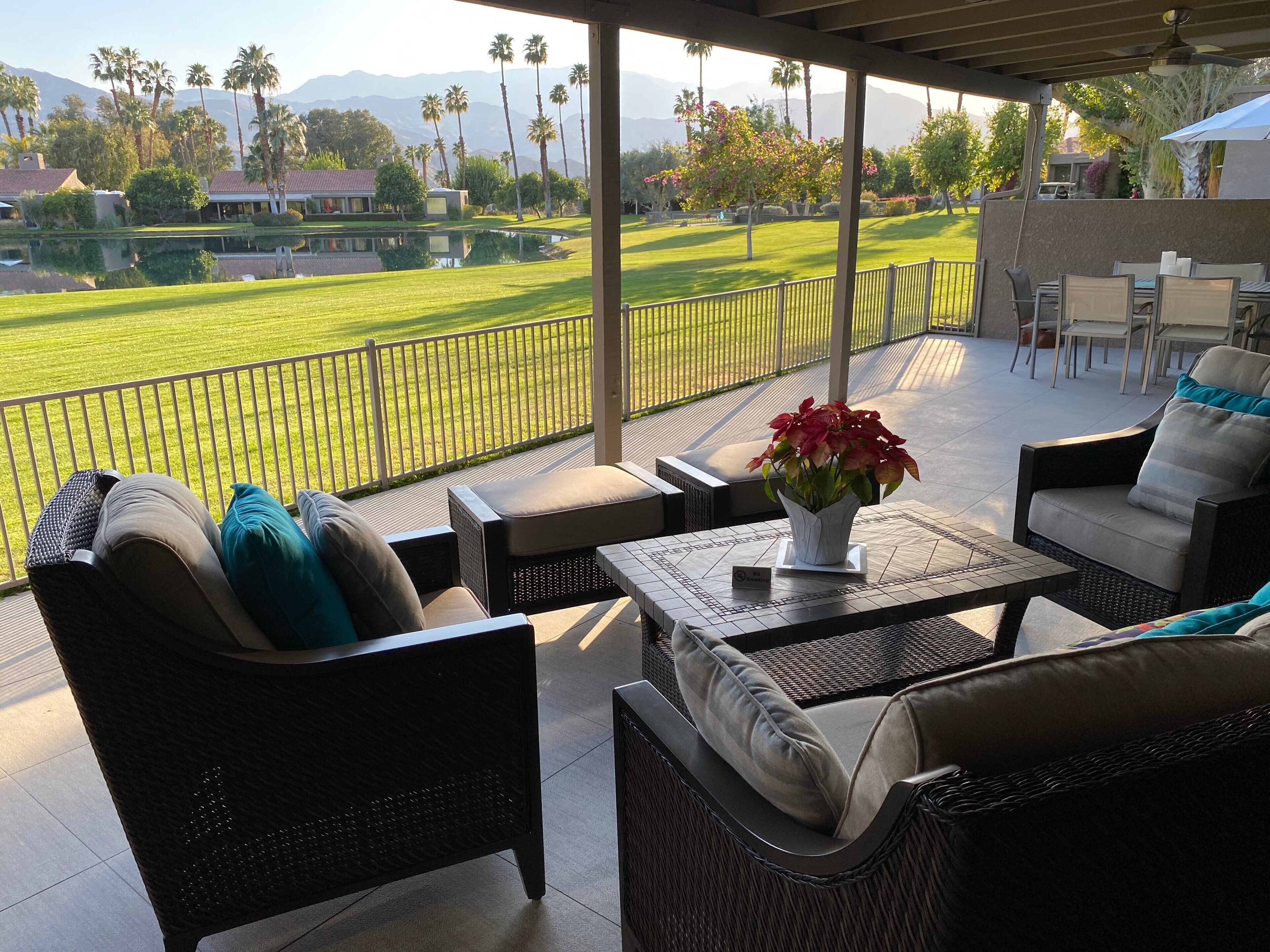 A covered outdoor seating area with wicker furniture, a table with a vase of flowers, and a view of a golf course and mountains.