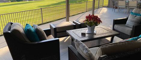 A covered outdoor seating area with wicker furniture, a table with a vase of flowers, and a view of a golf course and mountains.