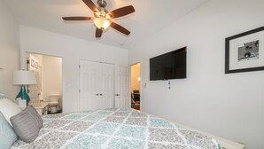 Un dormitorio con cama, ventilador de techo, televisor y puerta que da a otra habitación.