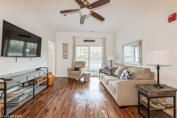 A living room with a wooden floor, a ceiling fan, a sofa, a coffee table, a TV, and a bookshelf.