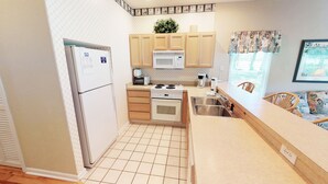 A compact kitchen with a refrigerator, microwave, stove, and sink. There is a window with a view of the outdoors, and a chair is visible in the corner. - Club Villas Unit 25A - Lower Level (Sunset Beach)