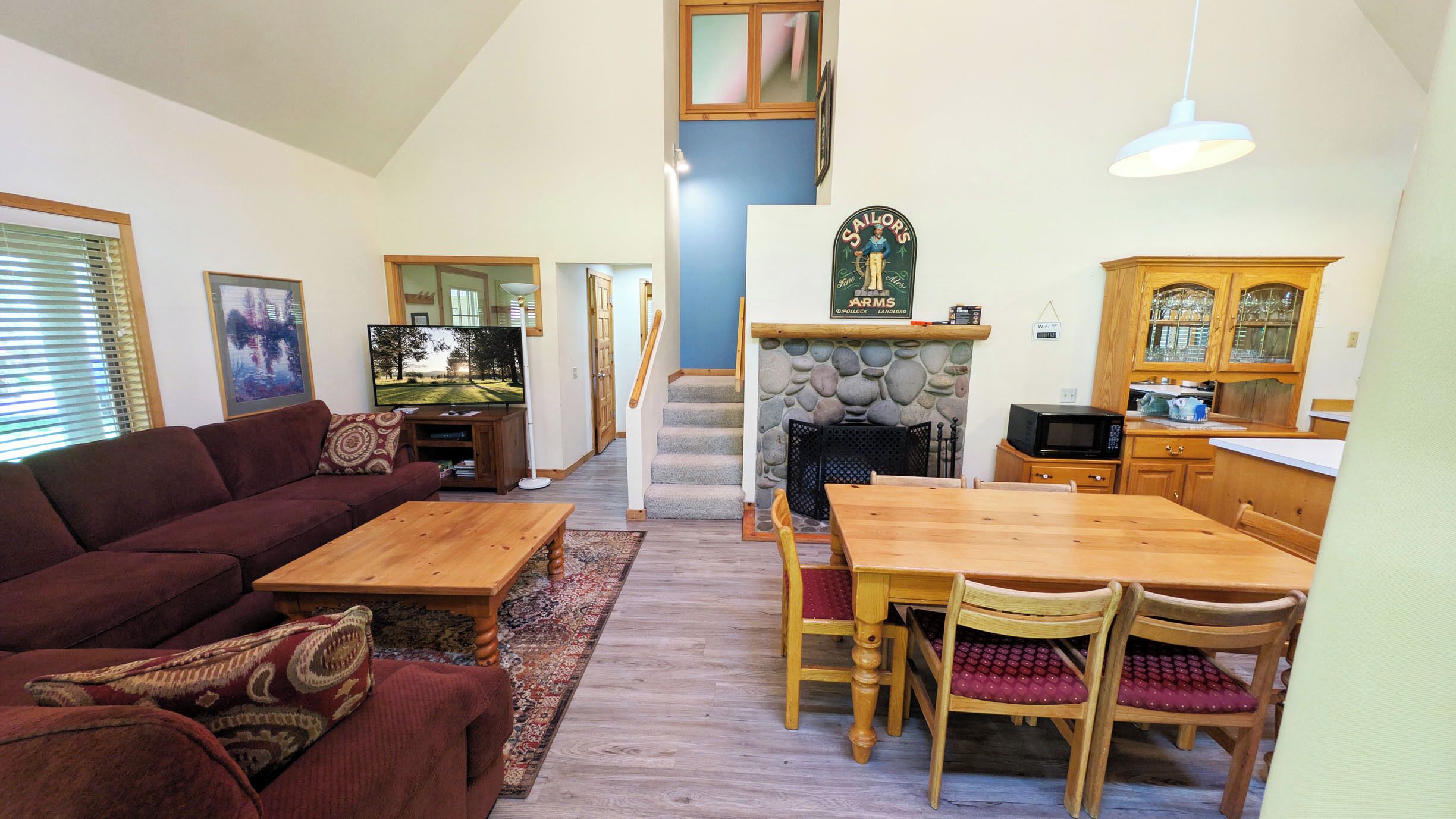 A spacious living room with a large sofa, a wooden coffee table, a dining table with chairs, a stone fireplace, a kitchen area with wooden cabinets, and a staircase leading to an upper level.