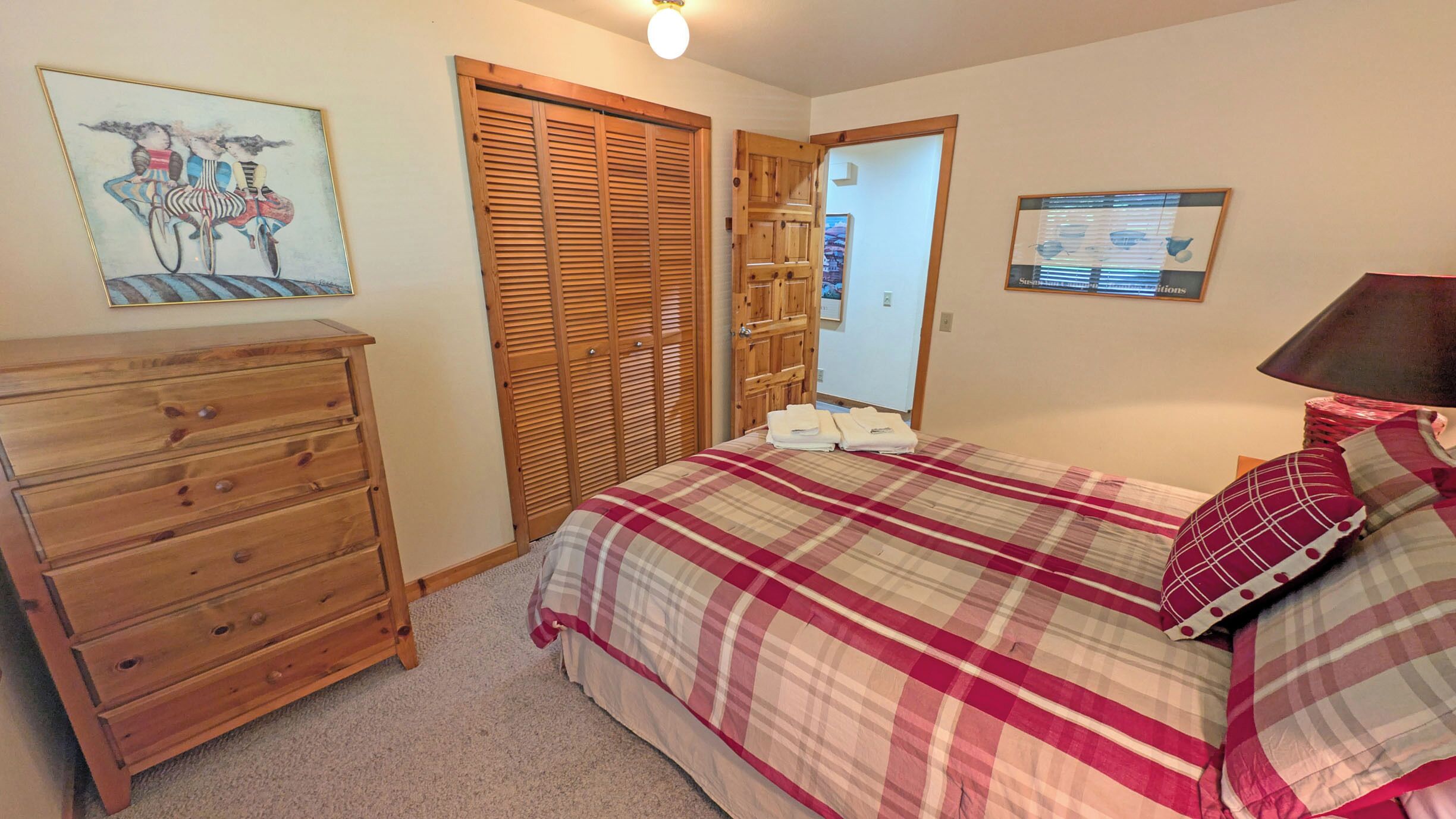 Une chambre avec une commode en bois, un lit recouvert d’une couverture à carreaux, une armoire en bois et un mur orné d’un tableau encadré et d’un tableau d’affichage.