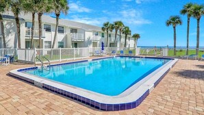 Une piscine rectangulaire bordée de carreaux bleus, entourée d’une clôture blanche, avec des palmiers en arrière-plan.