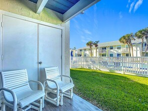 A covered outdoor seating area with white chairs and a door. - Colony Beach Club 146 - Cottage By The Sea (New Smyrna Beach)