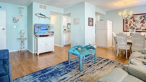 Un salon spacieux doté d’un parquet, d’un canapé bleu, d’une table basse turquoise et d’un coin repas avec des chaises blanches et un lustre.