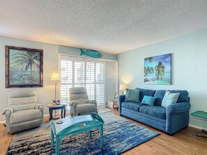 A living room with a blue couch, two armchairs, a coffee table, and a rug. There are two paintings on the walls, a window with blinds, and a door. - Colony Beach Club 146 - Cottage By The Sea (New Smyrna Beach)