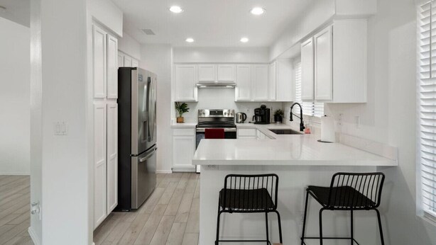 A modern kitchen with white cabinetry, a stainless steel refrigerator, a gas stove, and a sink. There are two black bar stools at the island. - City Escape in Goodyear w Pool Spring Training (Goodyear)