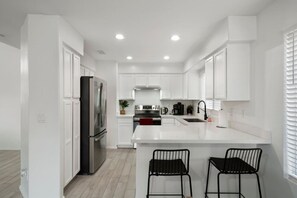 A modern kitchen with white cabinetry, a stainless steel refrigerator, a gas stove, and a sink. There are two black bar stools at the island.