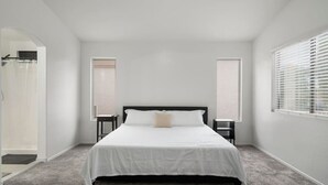 A bedroom with a bed, two nightstands, a window with blinds, and a door leading to a bathroom. - City Escape in Goodyear w Pool Spring Training (Goodyear)