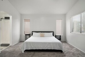 A bedroom with a bed, two nightstands, a window with blinds, and a door leading to a bathroom.