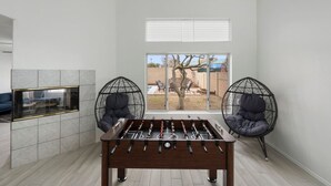 A modern room with a foosball table, two egg chairs, a glass fireplace, and a large window with a view of an outdoor area. - City Escape in Goodyear w Pool Spring Training (Goodyear)