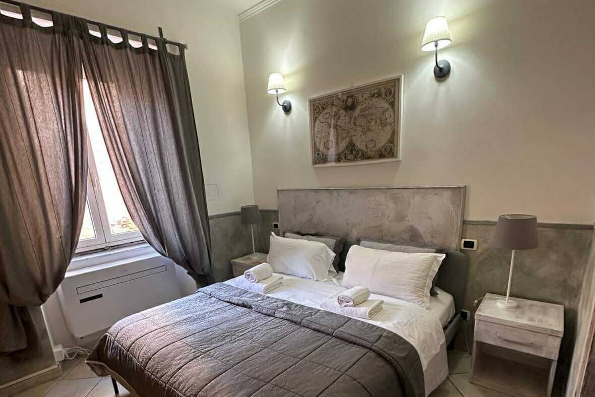 A bedroom with a bed, two bedside tables, a window with curtains, and a wall-mounted light fixture.