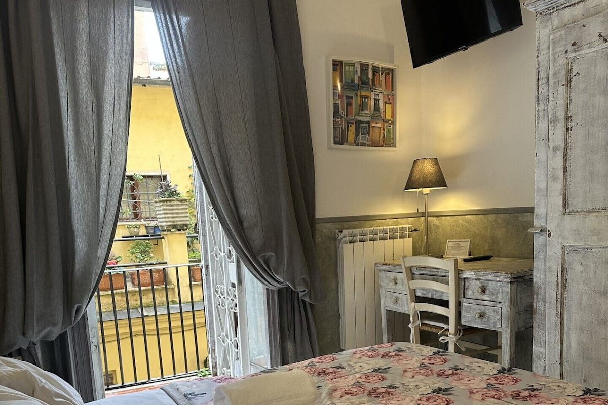 A bedroom with a bed, a desk, a chair, a TV, and a window with curtains.