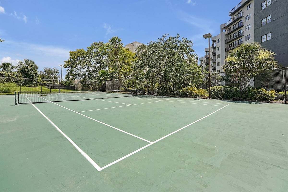 An outdoor tennis court with a net, surrounded by trees and buildings.