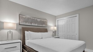 Un dormitorio ordenado con una cama grande, dos mesitas de noche con lámparas, una puerta y una obra de arte colgada en la pared.