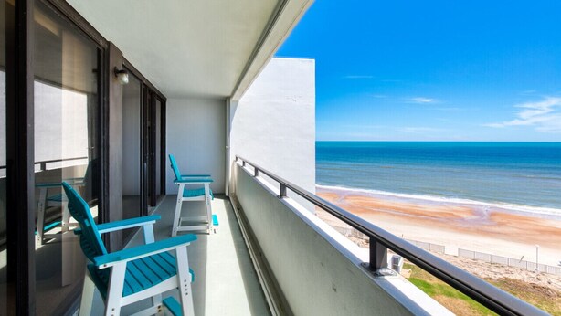 Varanda com duas cadeiras turquesa e uma mesa, com vista para uma praia e céu azul cristalino.