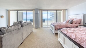 Quarto com uma grande porta de correr de vidro que dá para uma varanda, um sofá cinzento e duas camas com roupa de cama rosa.