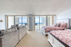 A bedroom with a large sliding glass door leading to a balcony, a grey sofa, and a bed with pink bedding. - Aliki Atrium Beachfront Condo (Ormond Beach)