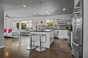 A modern kitchen with a central island, stainless steel appliances, and a dining area with bar stools. - Andreas (Palm Springs)