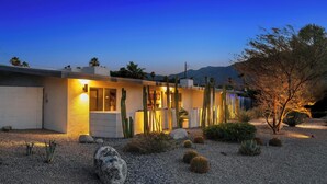 Une maison de style désertique, avec une allée en gravier, des cactus et une chaîne de montagnes en arrière-plan.