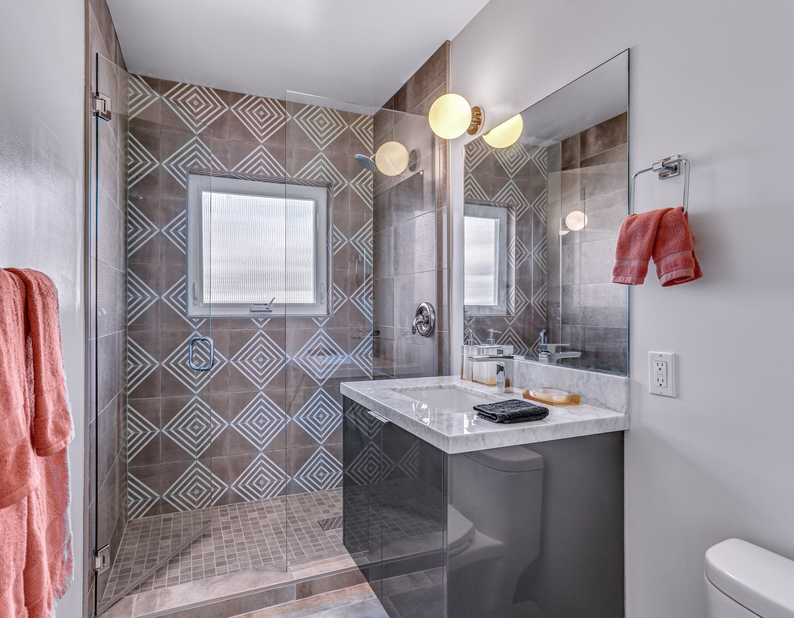 Un baño moderno con baldosas estampadas, cabina de ducha de cristal, tocador con espejo y inodoro.