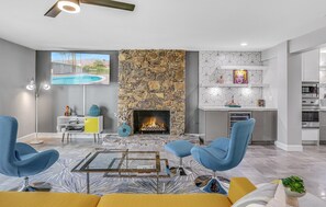 A modern living room with a stone fireplace, a pool view, and a kitchen area. - Farrell 2 (Palm Springs)