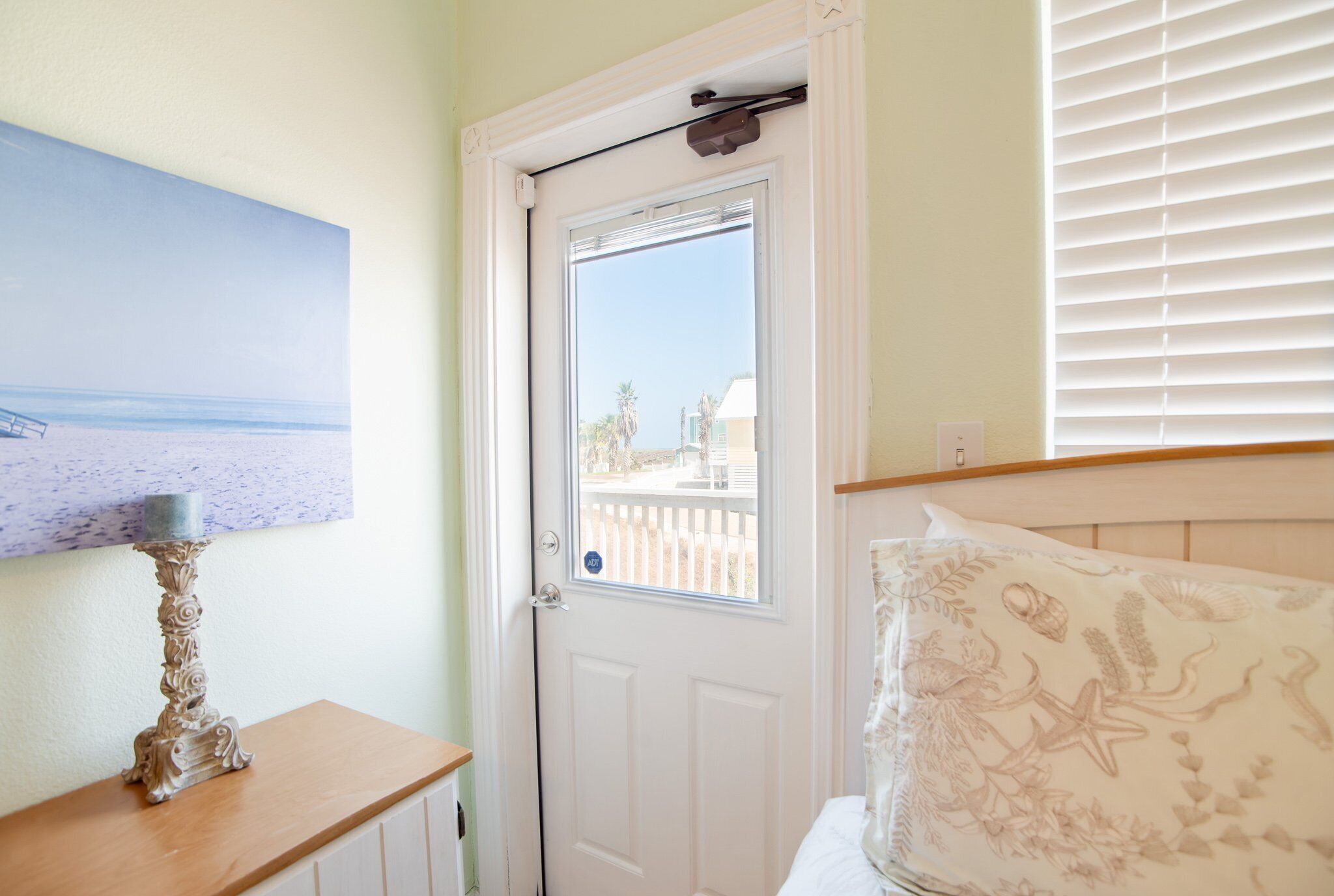 A room with a door, a small table with a candle, a bed with a floral pillow, and a painting of a beach scene.