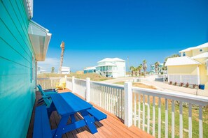Une terrasse en bois avec des tables de pique-nique bleues, une clôture blanche et des maisons de plage en arrière-plan.