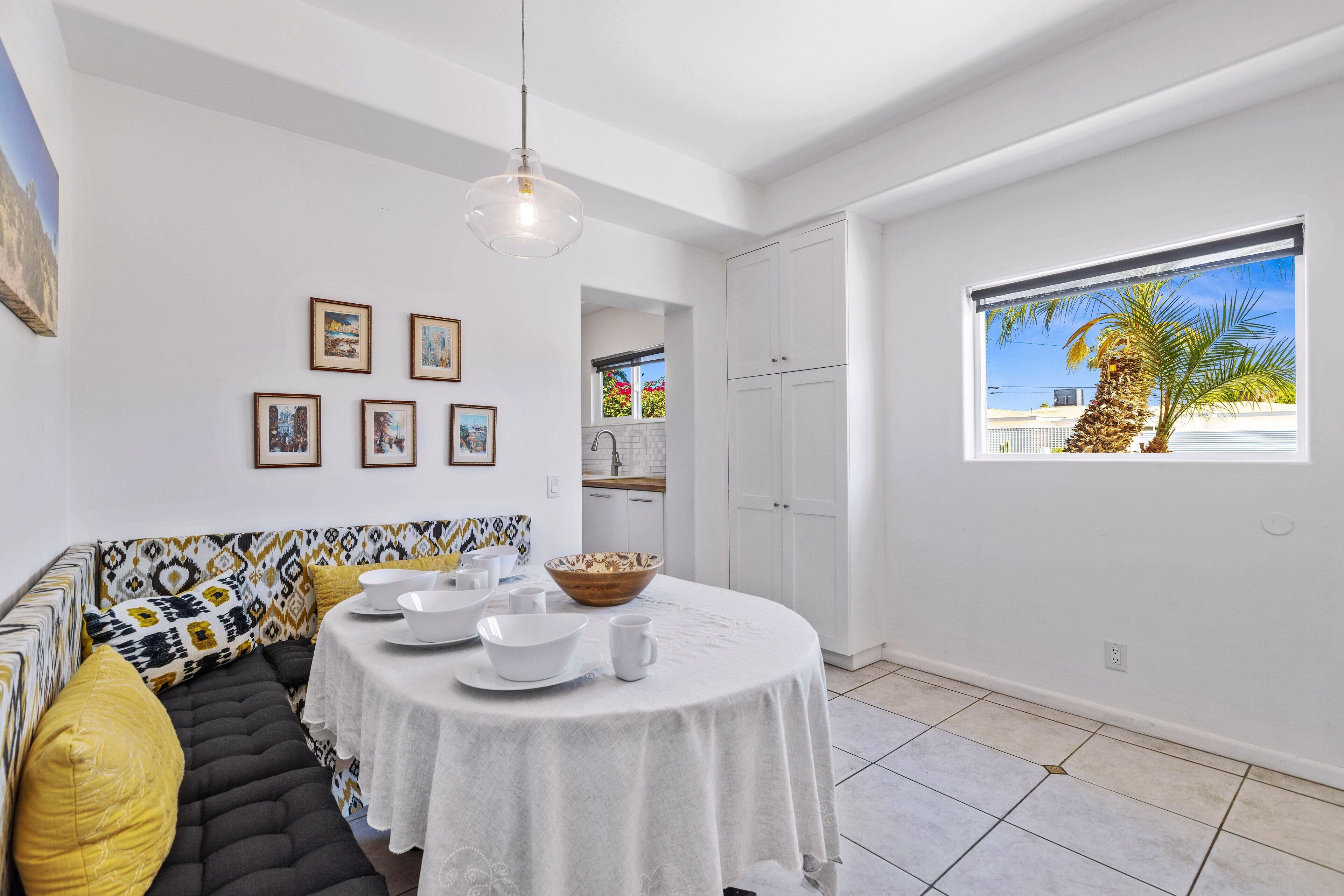 A modern kitchen with white cabinets, a dining area with a round table, and a large window with a view of palm trees.