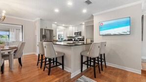 A modern kitchen with white cabinets, a central island with bar stools, a large flat-screen TV, and a dining area with a table and chairs. - Dockside Serenity (Palm Coast)