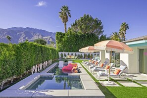 Zona de piscina con sillones de descanso y sombrillas, rodeada de vegetación y montañas al fondo.