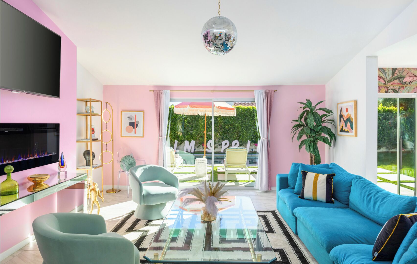 Un salon moderne aux couleurs rose et bleu, avec une table basse en verre, un canapé bleu et une grande fenêtre donnant sur un jardin.