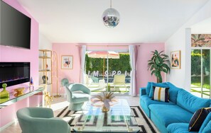 Una sala de estar moderna con una paleta de colores rosa y azul, una mesa de centro de vidrio, un sofá azul y un ventanal con vistas a un jardín.