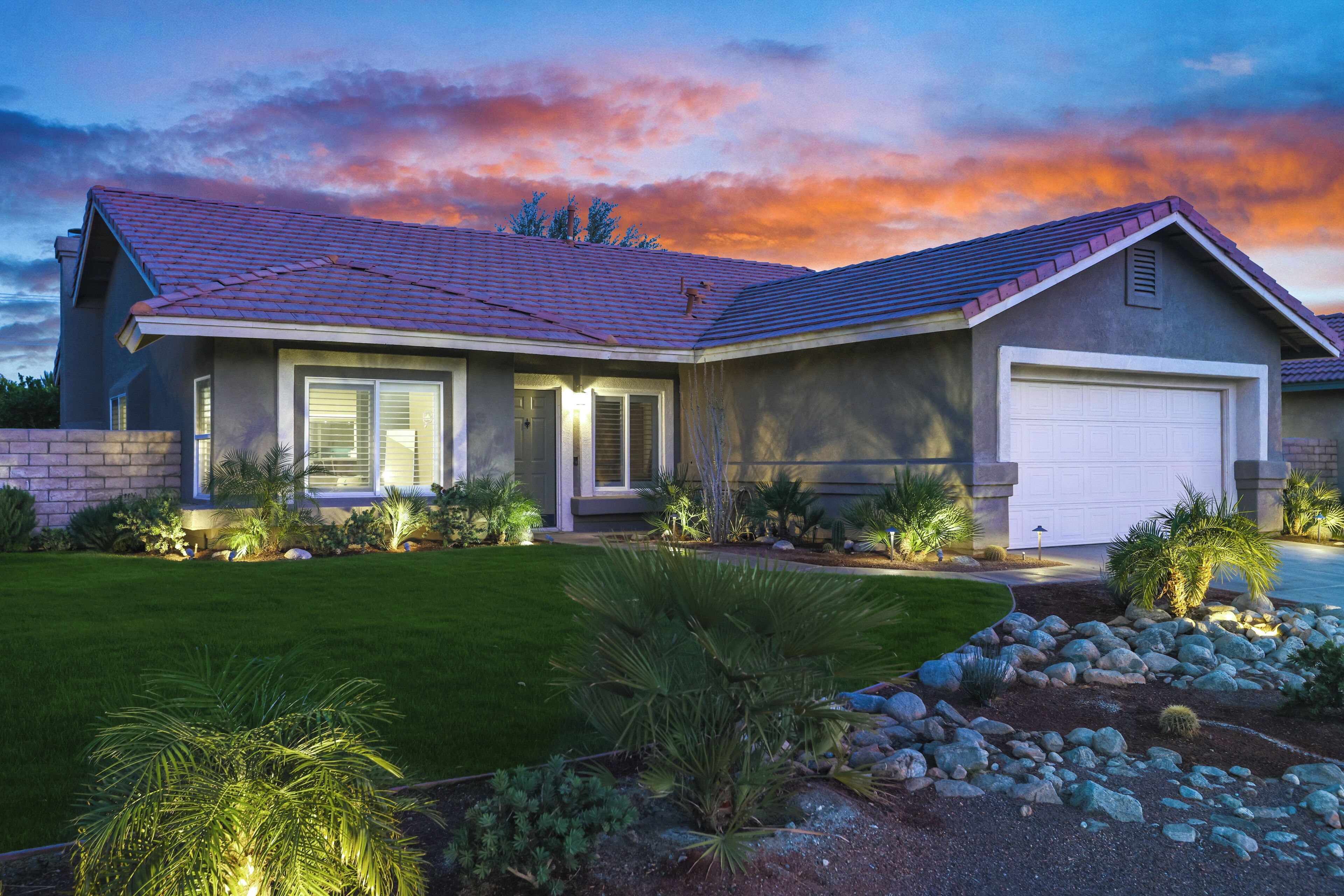A modern two-story house with a red-tiled roof, a white garage door, and a well-maintained front lawn with various plants and a rock garden.
