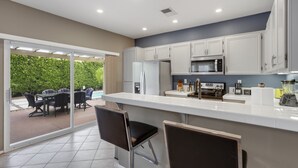 A modern kitchen with white cabinets, a stainless steel refrigerator, and a built-in microwave. There is a dining area with a table and chairs outside. - Shannon (Palm Springs)