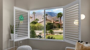 A room with a large window, a white wall, a small table, a chair, and a potted plant. - Shannon (Palm Springs)
