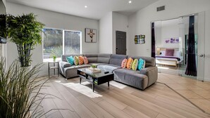 Un salon moderne avec un canapé d’angle gris, une table basse et une grande fenêtre équipée de stores.