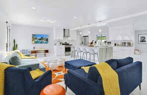 A modern living room with a sofa, coffee table, and chairs. There is a kitchen area with white cabinets and a dining area with bar stools. - Viminal (Palm Springs)
