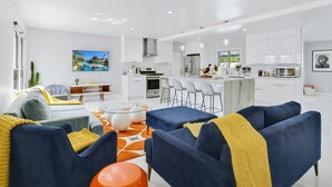 Een moderne woonkamer met een bank, salontafel en stoelen. Er is een keuken met witte kasten en een eettafel.
