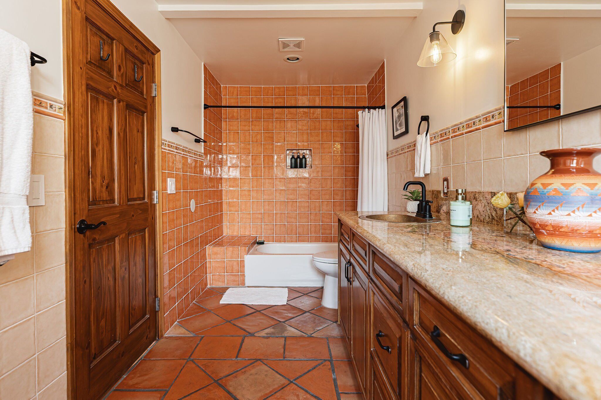 A bathroom with a wooden door, a shower, a toilet, and a vanity with a large vase.
