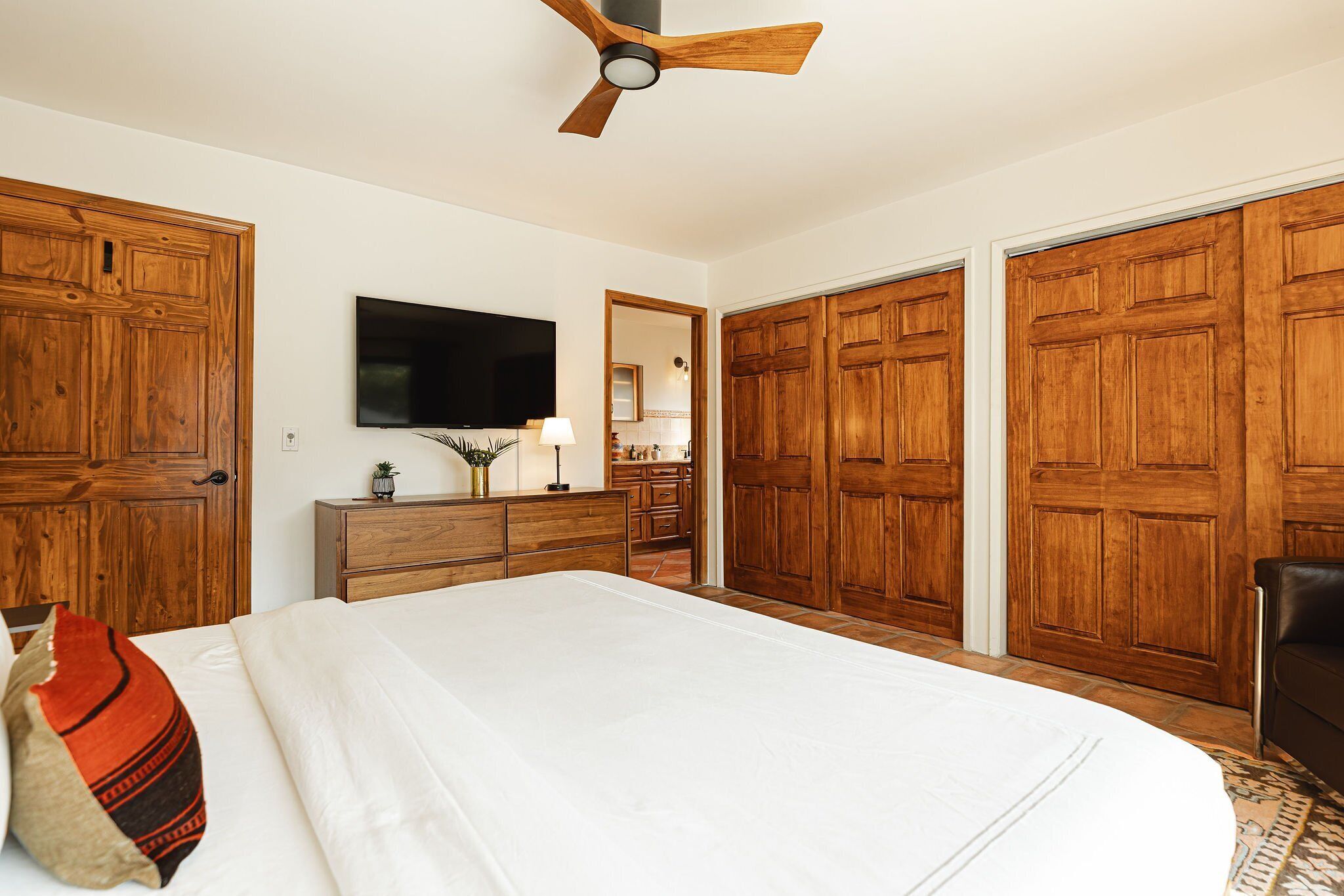 A bedroom with a large bed, wooden wardrobe, a ceiling fan, a TV, and a dresser.