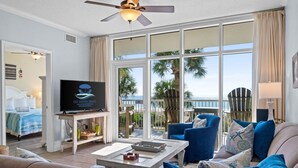 A spacious living room with a large TV, comfortable seating, a balcony with a view of the ocean, and a ceiling fan. - Waters Edge 205 - Melissa`s Happy Place (Fort Walton Beach)