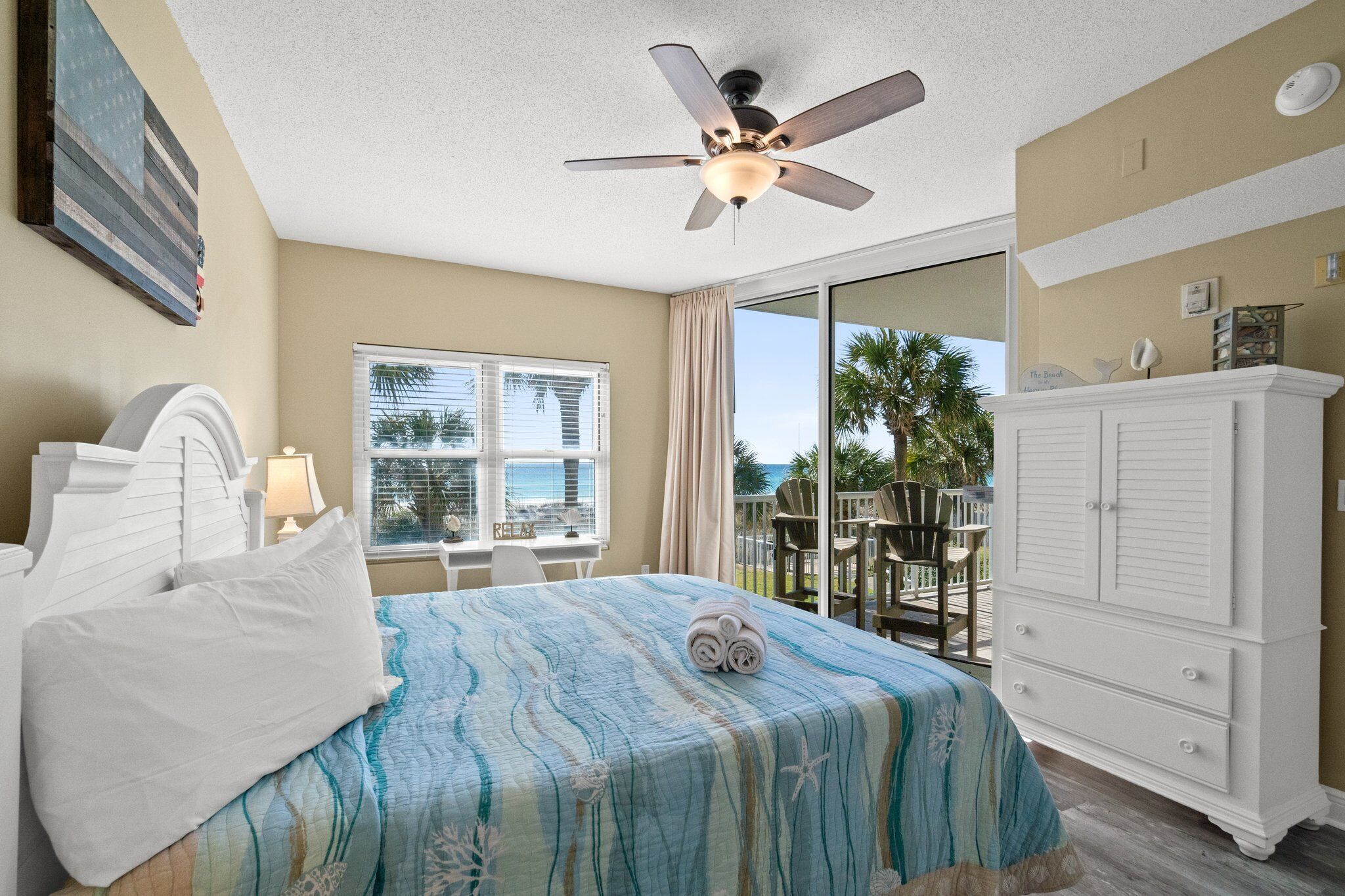 A hotel room with a bed, a ceiling fan, a window with a view of the ocean, a white dresser, and a balcony with chairs.