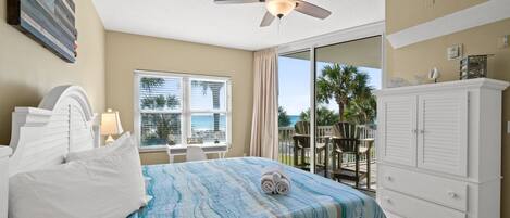 Une chambre d’hôtel avec un lit, un ventilateur de plafond, une fenêtre donnant sur l’océan, une commode blanche et un balcon avec des chaises.