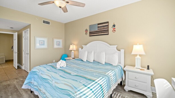 A bedroom with a bed, white nightstands, a ceiling fan, and wall decorations including a flag and a lantern. - Waters Edge 205 - Melissa`s Happy Place (Fort Walton Beach)