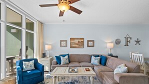 A living room with a blue armchair, beige sofa, and a coffee table. There are wall decorations, a ceiling fan, and a window with curtains. - Waters Edge 205 - Melissa`s Happy Place (Fort Walton Beach)