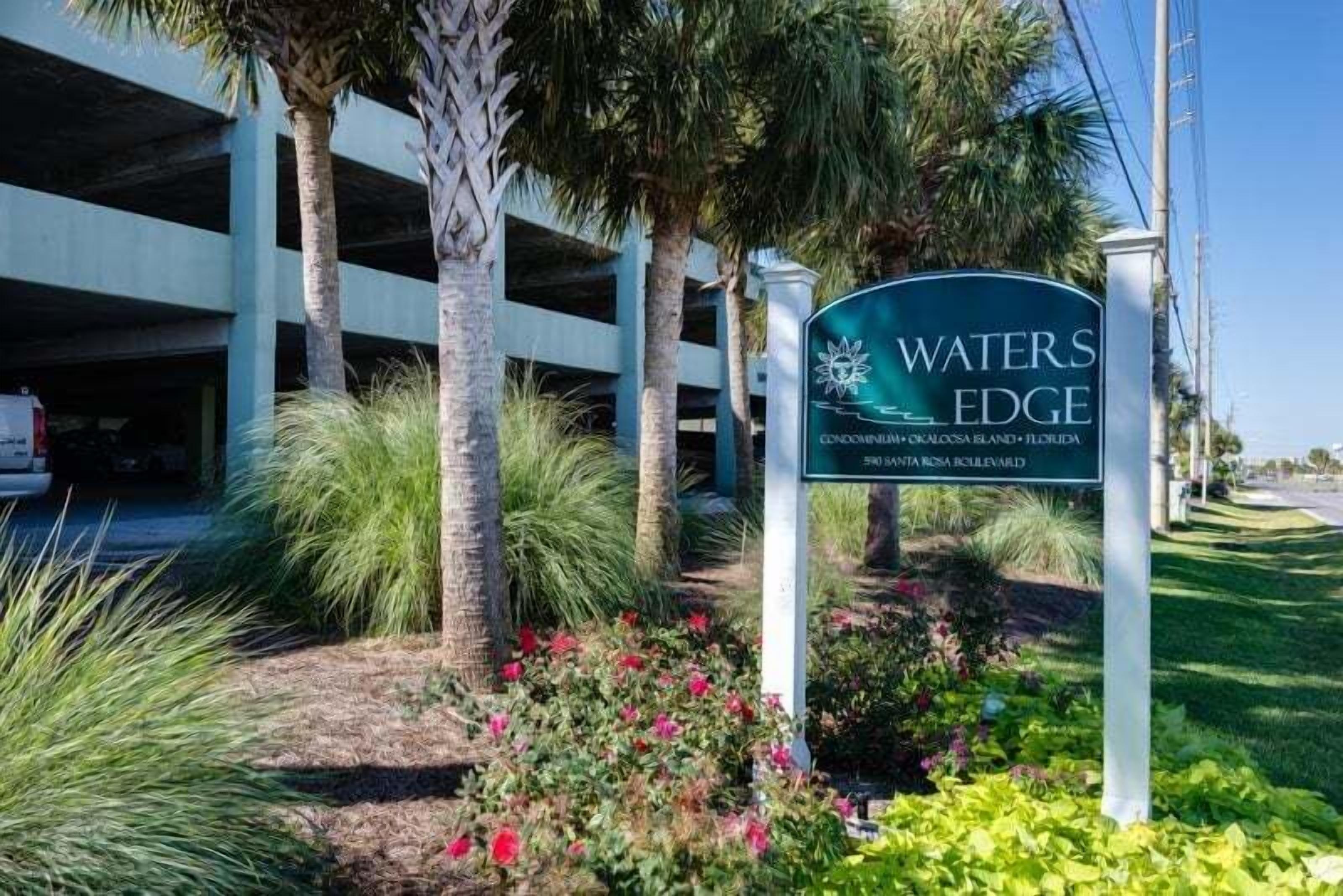 Naamplaatje voor 'WATERS EDGE' met uitzicht op een parkeergarage en een aangelegde omgeving.