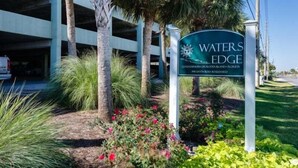 A sign for 'WATERS EDGE' with a view of a parking garage and landscaped area. - Waters Edge 404 - Okaloosa Dreamin' (Fort Walton Beach)