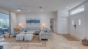 A spacious living room with a large sofa set, a coffee table, and a dining area. There is a large window providing natural light and a view of the outdoors. - Life's Porpoises - 29 Coconut Ct (Palm Coast)